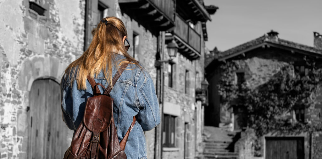 Jeune femme en couleur marchant avec une veste en jean et un sac à dos dans un village médiéval sur fond en noir et blanc.